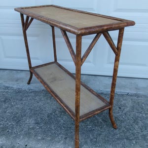 May include: A rectangular console table constructed from brown bamboo with a mottled finish. The table features two horizontal shelves, each with a flat, light tan woven surface, supported by four legs connected by diagonal bamboo bracing.