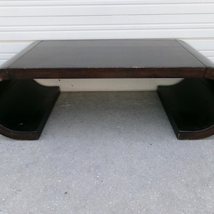 May include: A dark brown, curved coffee table with a rectangular top and rounded ends. The table has a unique, modern design with a dark finish. The table is approximately 48 inches long, 20 inches wide, and 16 inches high.