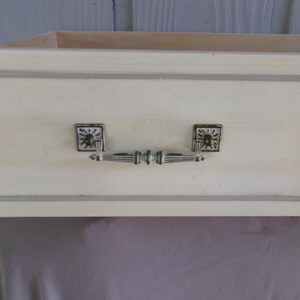 May include: A close-up of a drawer front with a cream-colored finish and a gray border. The drawer has two square metal pulls with a sunburst design and a decorative handle. The drawer is part of a larger piece of furniture.