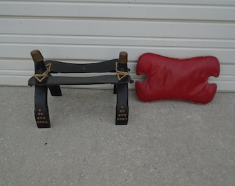 May include: A black wooden saddle stool with brass accents and a red leather cushion. The stool has a simple design and is perfect for adding a touch of rustic charm to any room.