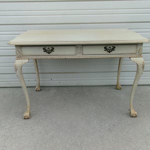 May include: A cream-colored wooden desk featuring a rectangular top, two drawers with ornate black metal pulls, and four curved cabriole legs that terminate in ball and claw feet. The apron is decorated with a carved rope-like trim detail.