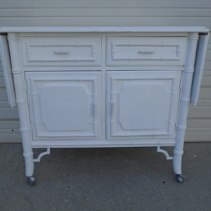 May include: White painted bamboo-style cabinet with two drawers and two doors. The cabinet has drop-leaf sides and sits on casters. The design includes bamboo-like detailing on the legs and drawer fronts.