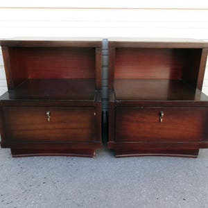 May include: Two matching dark wood nightstands. Each nightstand has a drawer with a keyhole and a shelf above. The nightstands have a curved base and a dark finish. The nightstands are made of wood.