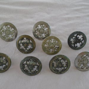 May include: A collection of ten antique metal drawer pulls. Each pull is circular with an openwork design featuring a central floral motif and an outer ring. The pulls have a tarnished, aged appearance, with variations in color.
