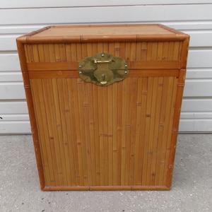 May include: A rectangular bamboo storage chest with a hinged lid and brass-colored clasp. The chest is constructed from vertical bamboo poles and has a natural, light brown finish. The clasp is ornate and adds a decorative element to the chest.