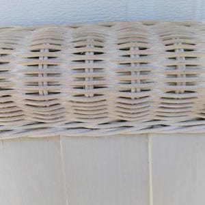May include: Close-up of a section of a white wicker chair. The image shows the woven texture of the wicker, with horizontal and vertical lines creating a grid pattern. The chair's frame is visible at the bottom, painted in a matching white.