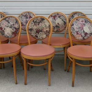 May include: A set of ten vintage wooden chairs. Each chair has a round, peach-colored seat and a floral-patterned backrest. The chairs have a light brown wooden frame.