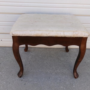 May include: A small, antique wooden stool with a cream-colored, patterned upholstered seat. The stool has four curved legs and a dark brown finish, suitable for a bedroom or living room.