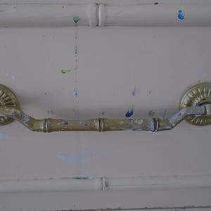 May include: A gold-toned drawer pull with a bamboo-style design. The pull has a curved shape and is attached to a white surface.