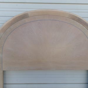 May include: A light-colored wooden headboard with an arched top. The headboard features a detailed design with a series of horizontal lines along the top edge and a subtle wood grain pattern.