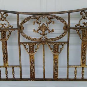 May include: Ornate, antique-style metal headboard with a gold finish. The headboard features an arched top, an oval center design with a fleur-de-lis, and decorative vertical supports with intricate scrollwork and baluster details.