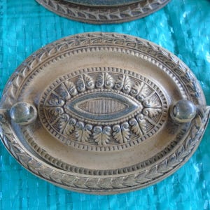 May include: Oval-shaped, antique-style drawer pulls with ornate detailing. The pulls are beige with darker accents and feature a central oval design with leaf and scroll patterns. Each pull has a rounded handle.