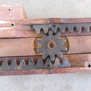 May include: A mechanical assembly featuring a central dark metal gear engaged with two parallel horizontal metal racks. The gear and racks are mounted onto a brown wooden frame with visible screw fasteners and signs of wear on the wood surface.