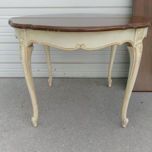 May include: A round table with a dark brown wooden top and a cream-colored base. The table features four curved cabriole legs with decorative carvings at the top. The apron of the table has a scalloped edge design with subtle gold-toned accents.