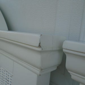 May include: Close-up of a section of a white painted radiator cover. The cover has a decorative design and is made of wood. The paint is chipped in some areas. The background is a textured white wall.