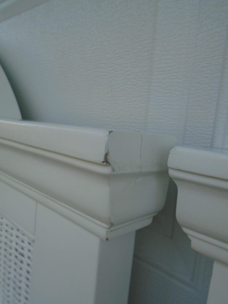 May include: Close-up of a section of a white painted radiator cover. The cover has a decorative design and is made of wood. The paint is chipped in some areas. The background is a textured white wall.