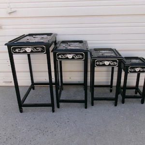 May include: Set of four black nesting tables with square tops and slender legs. Each table features a decorative white floral and scroll design on the front. The tables vary in height, creating a tiered display.