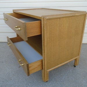 Puede incluir: Una mesita de noche de madera marr&oacute;n claro con dos cajones, ambos parcialmente abiertos. Los cajones tienen tiradores dorados. La mesita de noche est&aacute; sobre ruedas peque&ntilde;as y tiene una parte superior rectangular.