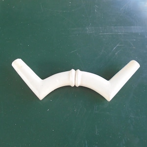 May include: A white, curved furniture handle with a central connecting piece and angled ends. The handle is made of a solid, opaque material and is set against a dark green background. The design is symmetrical and appears to be a decorative element.