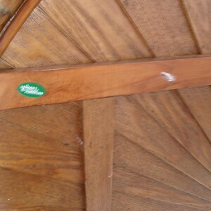 May include: Close-up of a wooden gazebo roof with a fan-like design. The wood has a warm, natural tone with visible grain. A green oval sticker with white text is affixed to a horizontal beam. The gazebo's structure is made of wood.