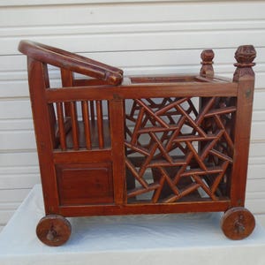 May include: A vintage wooden doll carriage with intricate geometric cutouts and small wooden wheels. The carriage features a curved handle and decorative finials. The wood has a warm, reddish-brown tone, suggesting age and craftsmanship.