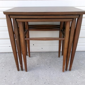 May include: Set of three nesting tables in a warm brown finish. Each table has a square top and tapered legs, designed to fit neatly together when not in use. The tables are made of wood.