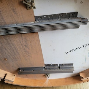 May include: Underside of a round wooden table with a light brown and white surface. Metal hardware and wooden supports are visible. The table has the text "H-6025-752" and the number "103" written on it.