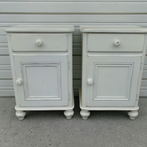 May include: A pair of matching off-white bedside tables, each featuring a single top drawer with a round knob and a lower cabinet door with a matching round knob. The furniture pieces have a distressed finish and rest on four small bun feet.