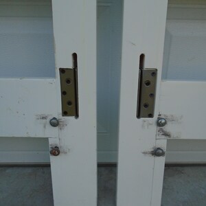 May include: Two white painted wooden door frames with metal hinges and bolts. The hinges are attached to the frame with screws. The frames are slightly weathered and show signs of use. The image is a close-up of the door frames.