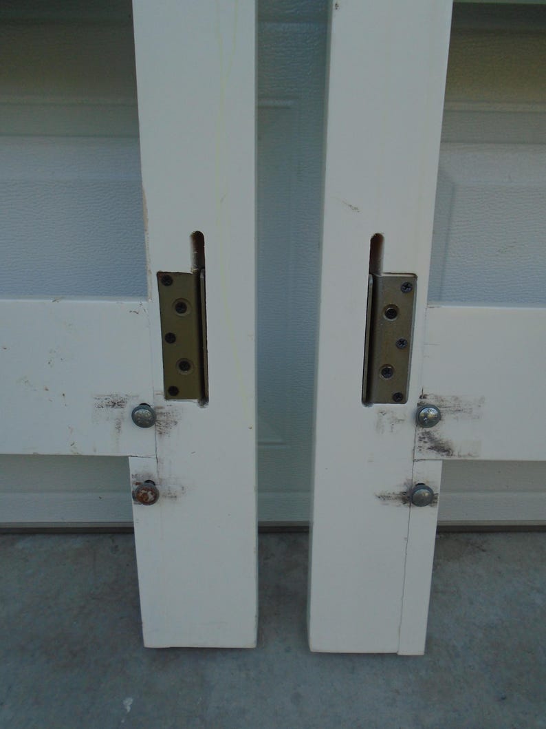 May include: Two white painted wooden door frames with metal hinges and bolts. The hinges are attached to the frame with screws. The frames are slightly weathered and show signs of use. The image is a close-up of the door frames.