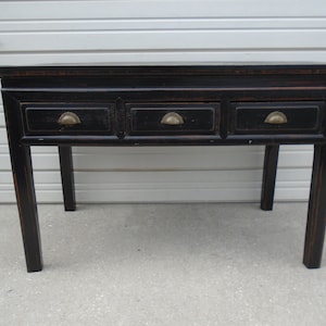 May include: A black wooden console table with three drawers and brass handles. The table has a distressed finish and sits on four legs.