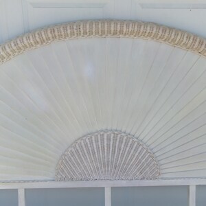 May include: A white, fan-shaped headboard with a woven wicker trim and a half-circle woven wicker design at the bottom. The headboard has a light, airy aesthetic, perfect for a bedroom or guest room. The headboard is made of wood and wicker.