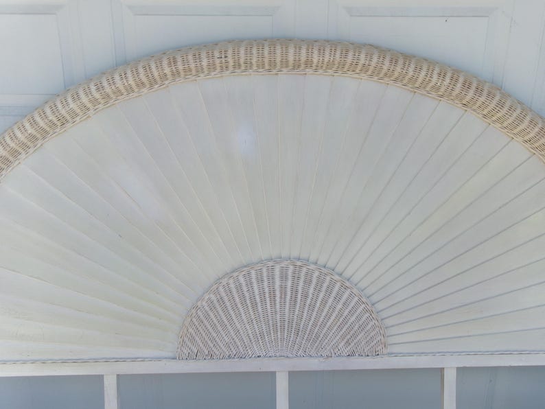 May include: A white, fan-shaped headboard with a woven wicker trim and a half-circle woven wicker design at the bottom. The headboard has a light, airy aesthetic, perfect for a bedroom or guest room. The headboard is made of wood and wicker.
