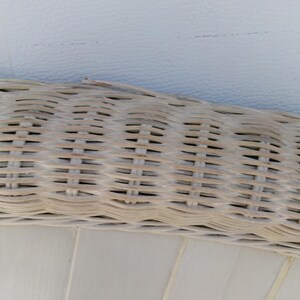 May include: Close-up of a section of a light-colored wicker weave, possibly from a chair or basket. The weave is tightly constructed with a natural, textured appearance. The background is a smooth, off-white surface.