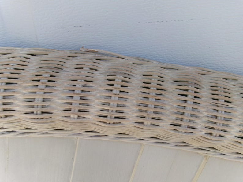 May include: Close-up of a section of a light-colored wicker weave, possibly from a chair or basket. The weave is tightly constructed with a natural, textured appearance. The background is a smooth, off-white surface.