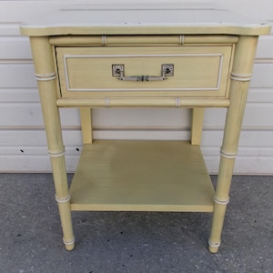 May include: A pale yellow side table with a single drawer and a lower shelf. The table has a bamboo-style design with white accents and a rectangular top. The drawer features a silver handle and decorative corner details.