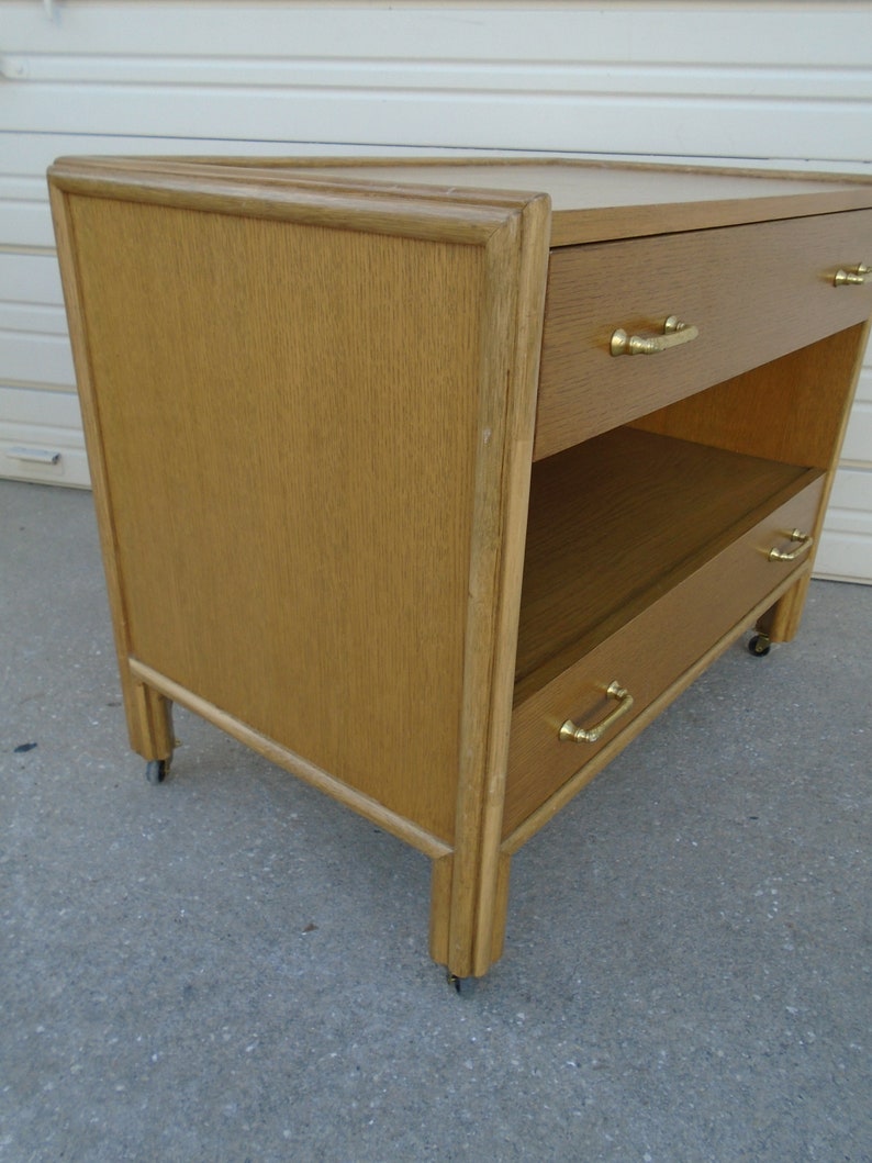 Puede incluir: Una mesita de noche de madera marr&oacute;n claro con dos cajones y un estante. La mesita de noche tiene tiradores de color lat&oacute;n y se apoya sobre peque&ntilde;as ruedas. La veta de la madera es visible y la pieza tiene un dise&ntilde;o moderno de mediados de siglo.