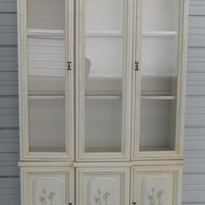 May include: Off-white wooden cabinet with three glass-door upper sections and three solid-door lower sections. The cabinet has a bamboo-style frame with green accents and decorative floral designs on the lower doors. The cabinet is designed for storage.