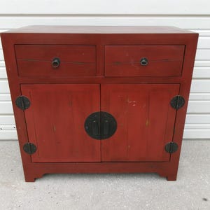 May include: A rectangular red wooden cabinet featuring two top drawers with circular metal pulls and two lower cabinet doors. The doors are secured with a large central circular metal plate and smaller circular hinges, showcasing a distressed finish.