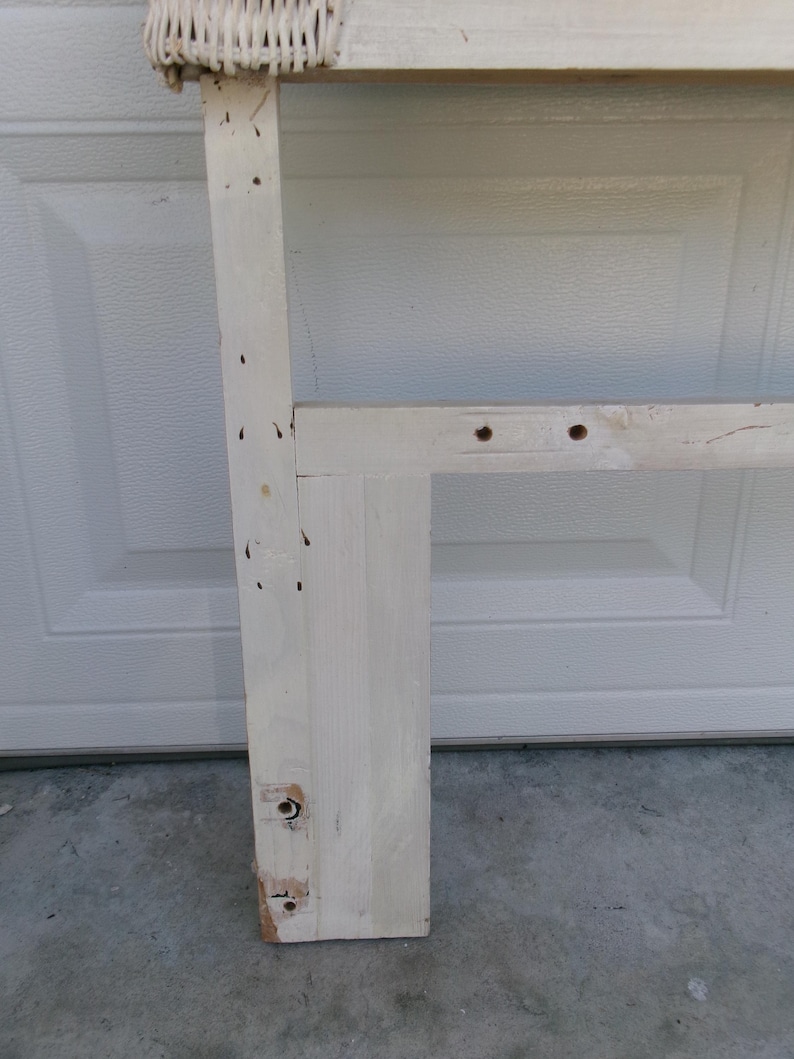 May include: A section of a white-painted wooden headboard frame with a woven wicker top. The frame has visible screw holes and some wear. The headboard is designed for a bed.