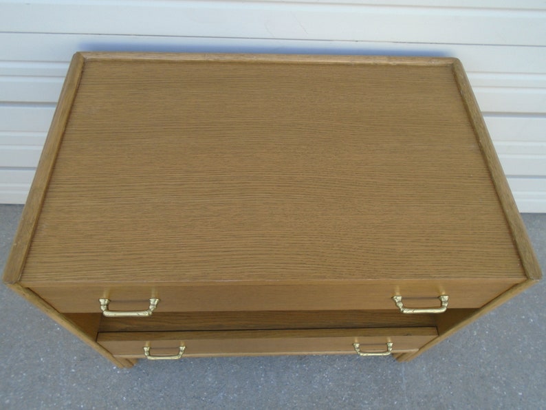 Puede incluir: Una c&oacute;moda de madera marr&oacute;n claro con dos cajones y tiradores dorados. La parte superior de la c&oacute;moda tiene un borde ligeramente elevado. La veta de la madera es visible.