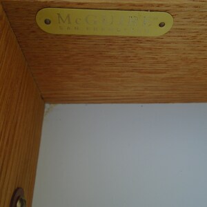 Puede incluir: Primer plano de una esquina de mueble de madera con una etiqueta McGuire de lat&oacute;n. La etiqueta es rectangular con bordes redondeados y el texto "McGUIRE SAN FRANCISCO" en una fuente serif. La veta de la madera es visible.