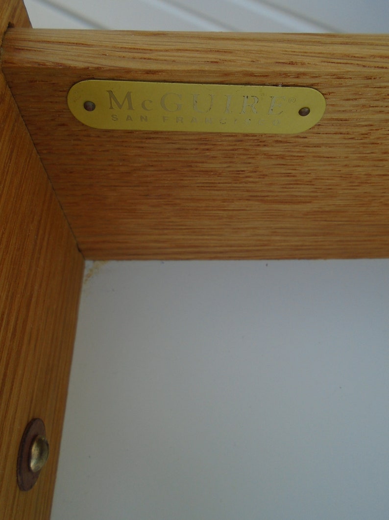 Puede incluir: Primer plano de una esquina de mueble de madera con una etiqueta McGuire de lat&oacute;n. La etiqueta es rectangular con bordes redondeados y el texto "McGUIRE SAN FRANCISCO" en una fuente serif. La veta de la madera es visible.