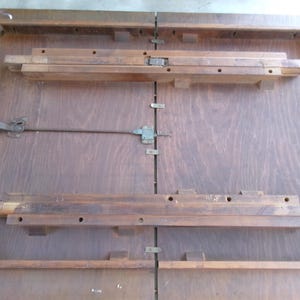 May include: A wooden table with a dark brown finish, featuring a folding mechanism. The table has a rectangular shape with a central seam and wooden supports. A metal rod and handle are visible, likely for opening and closing the table.