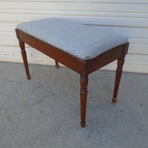 May include: A wooden piano bench with a rectangular seat covered in a light blue patterned fabric. The bench has four turned legs and a dark brown finish. The bench is designed for seating.