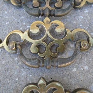 May include: Set of three ornate brass drawer pulls with a decorative scroll design. The pulls have a traditional style and are in a worn, antique finish.