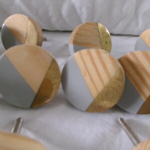 May include: A collection of round cabinet knobs with a modern design. Each knob features a combination of natural wood, gray paint, and gold-toned metal accents. The knobs are arranged on a white surface.