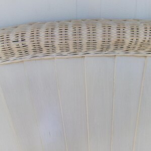 May include: Close-up of a section of a white wicker chair back. The top edge features a woven wicker design in a light beige color. The lower portion has vertical, flat, white panels, creating a fan-like pattern.