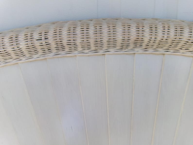 May include: Close-up of a section of a white wicker chair back. The top edge features a woven wicker design in a light beige color. The lower portion has vertical, flat, white panels, creating a fan-like pattern.