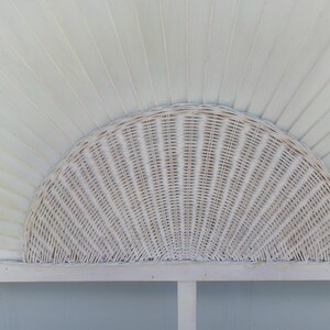 May include: A white, semi-circular wicker headboard with a woven pattern. The headboard is mounted on a white frame and set against a backdrop of white, slatted wood. The design evokes a coastal or bohemian aesthetic.